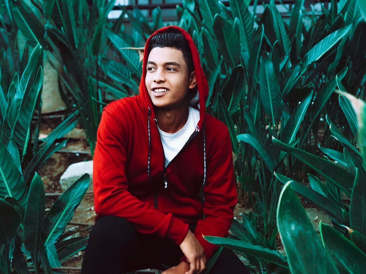 Portrait of a smiling young man in a red hoodie surrounded by lush green plants outdoors.