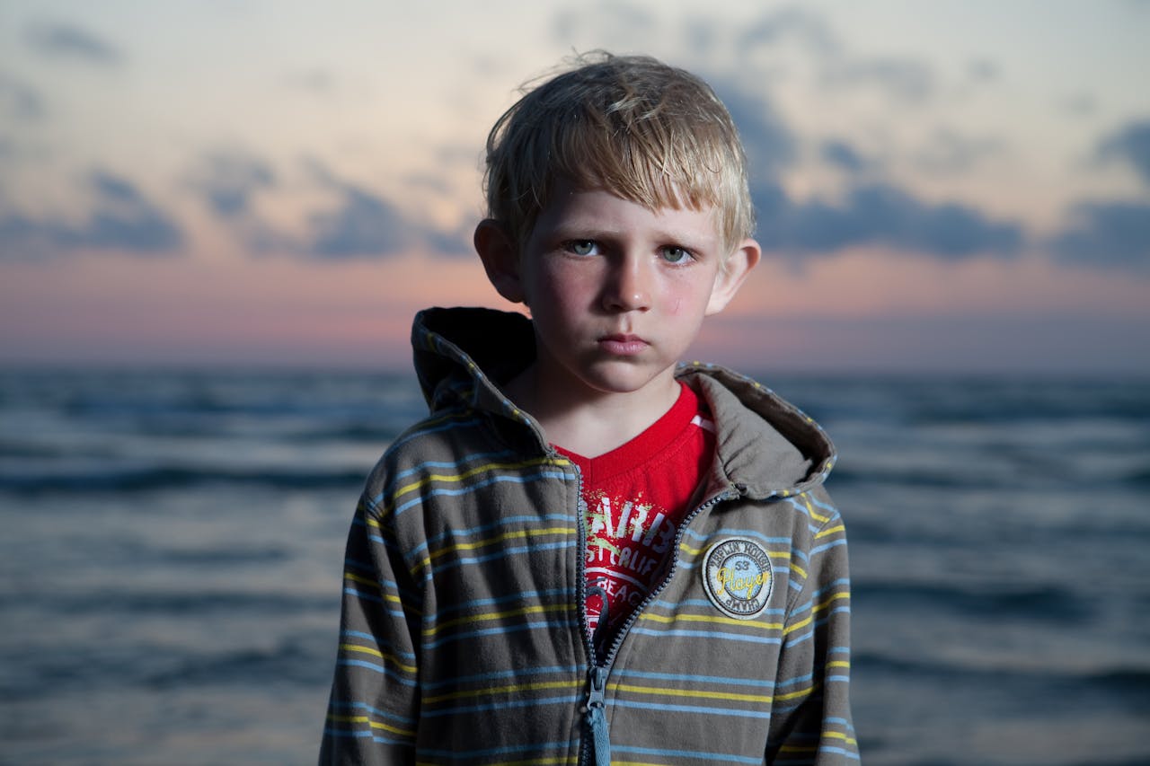 A blond boy in a hoodie stares seriously at the camera with a sunset over the sea behind him.